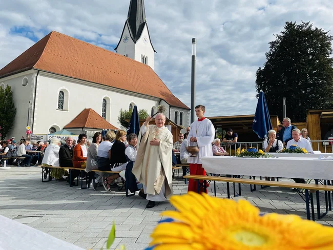 Ein Priester läuft mit Ministranten über den neuen Dorfplatz und weiht diesen mit Weihwasser.
