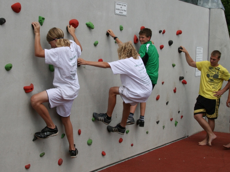 Drei Kinder klettern an einer Indoor-Boulder-Wand. Ein Erwachsener passt auf.